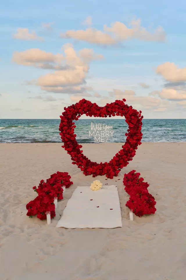 A deep red rose heart arch with a neon Will You Marry Me sign on the beach, a white mat and a bouquet of cream roses laid out in front of it