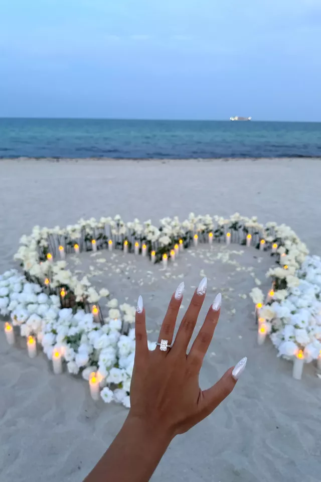 A newly engaged woman holds up her hand to show the ring, a large heart of white roses and lit candles on the sand behind her