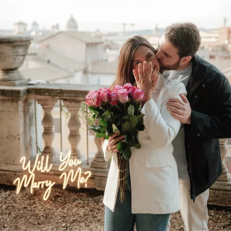 A newly engaged couple on a Rome rooftop terrace with pink roses and a neon Will You Marry Me sign, the dome of St Peter's behind them
