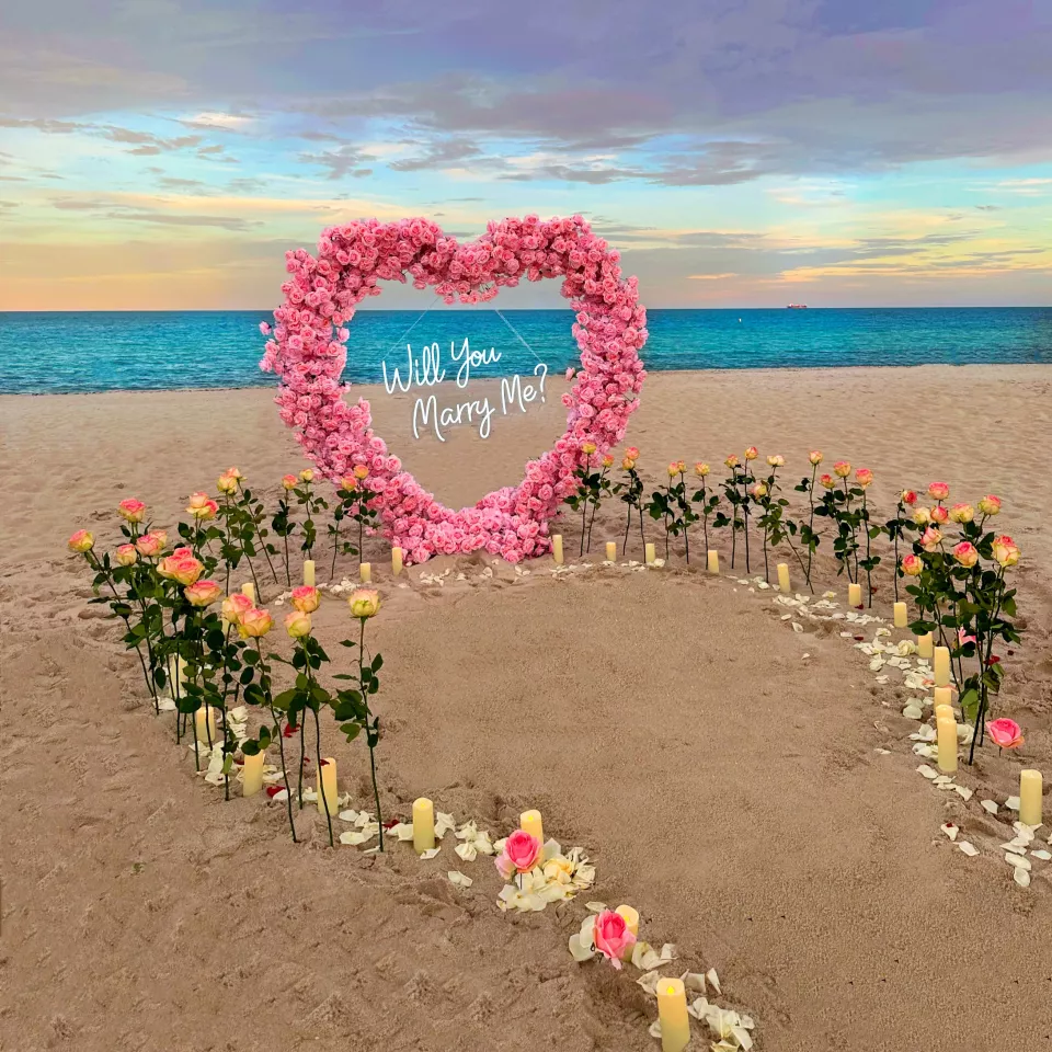 A pink rose heart arch with a neon Will You Marry Me sign on the sand, a path of roses and candles leading up to it and turquoise water behind